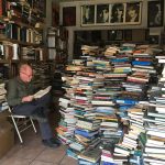 Pic: Man sitting reading in bookstore facing feet-high stacks of books